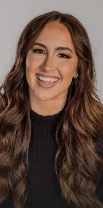 Smiling woman with long wavy brown hair wearing a black top against a gray background.